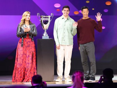 Three people standing on a stage, facing the audience, with a large silver cup trophy.