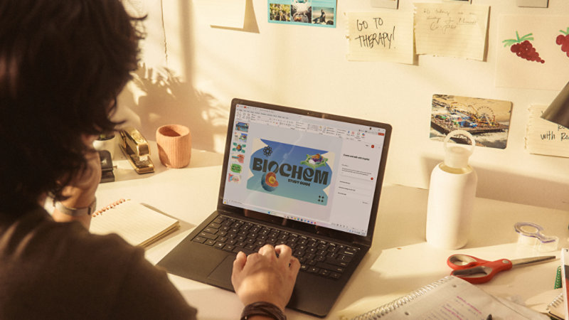 A person using a Surface laptop at a desk filled with notebooks, handwritten notes, and desk accessories.
