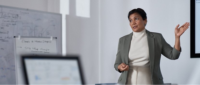 Femme professionnelle faisant une présentation dans une salle de réunion avec un tableau blanc et un ordinateur en arrière-plan.