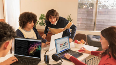 A group of students collaborate around a table with laptops, headphones, and notebooks.