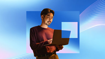 A man, standing, holding a laptop in his left hand, and using his right hand to type and operate the trackpad.