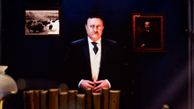 An image of United States president Theodore Roosevelt Standing behind a desk, resting his hands on a row of large books.