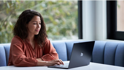 A woman sits by a large window using a laptop on a blue bench.