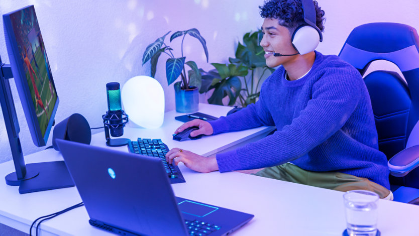 A person plays a game at a desk with a laptop, monitor, headset, and microphone.