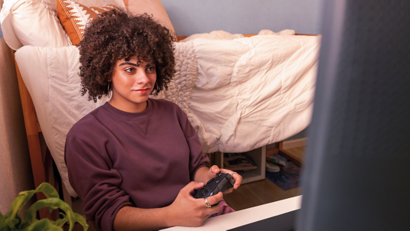 A person relaxes on a bed while holding a game controller and looking toward a large screen.