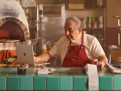 Man sitting at a countertop in a restaurant kitchen using a Surface Laptop and a calculator.