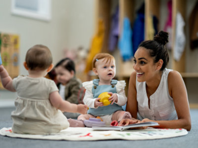 A caretaker smiles as she reads a picture book to two babies.