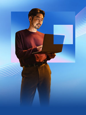 A man, standing, holding a laptop in his left hand, and using his right hand to type and operate the trackpad.