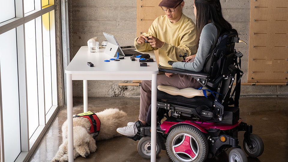 Человек, передвигающийся в инвалидной коляске, тестирует адаптивные аксессуары Майкрософт вместе с коллегой.