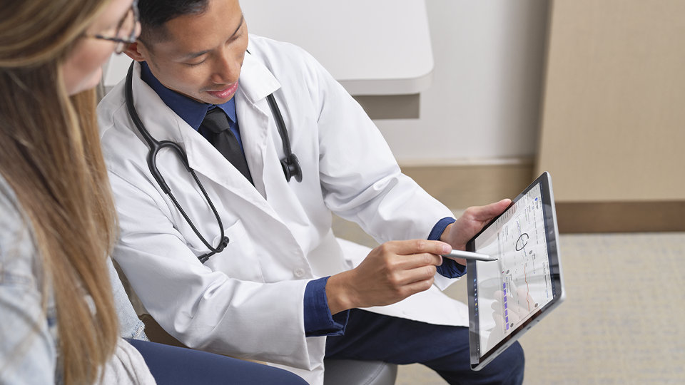 法人向け Surface ペンを使用して、法人向け Surface Go 4 のタッチスクリーンを操作しながら患者と話をする医師。