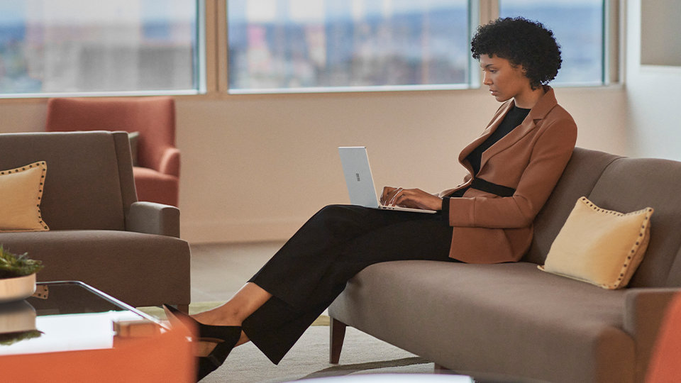 A person sits on couch in an office setting working on a Surface device.