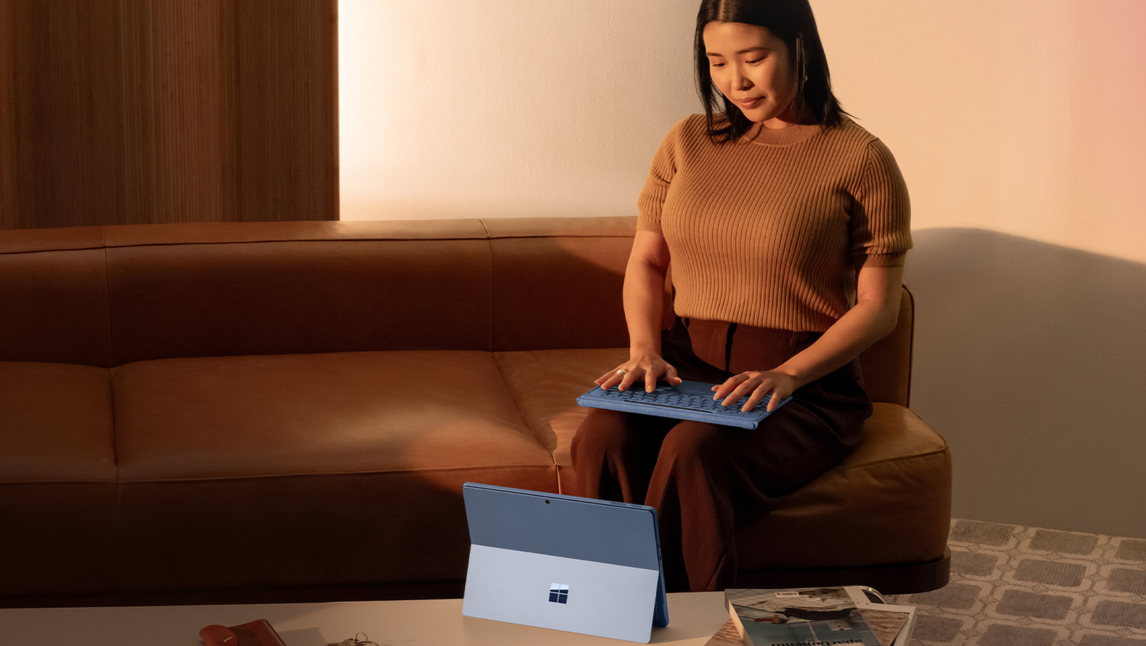 Eine Person benutzt ein nicht angebrachtes Surface Pro Flex Keyboard mit Slim Pen, das auf ihrem Schoß liegt, um auf einem Surface zu tippen, das auf einem Tisch aufgestellt ist. 