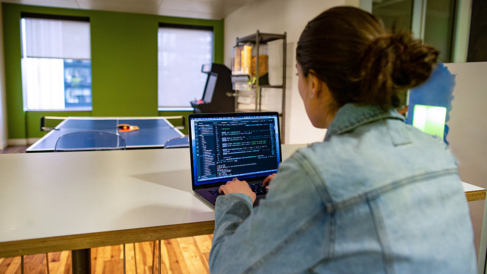 A person reviews code on a laptop while sitting near a ping pong table.