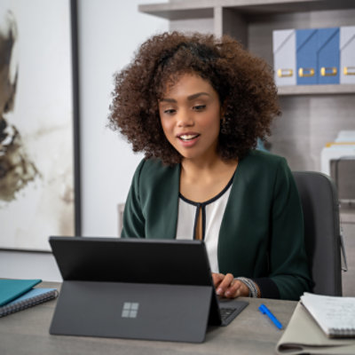Eine Frau, die in einem Büro sitzt und mit einem Microsoft Surface Pro arbeitet und lächelt. Notizbücher sind auf der Seite gelegt