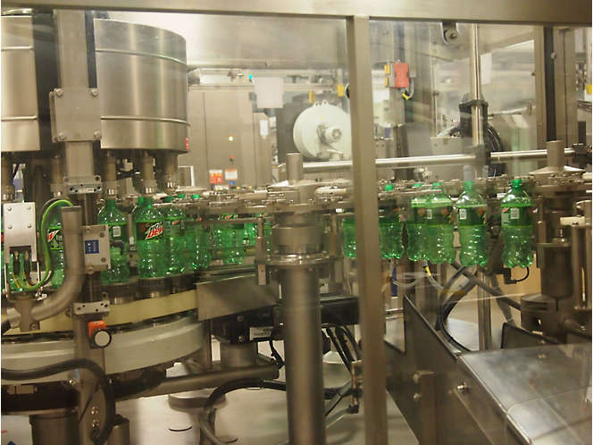 Išpilstymo į butelius gamykla, kurioje žali buteliai pripildomi ir uždaromi automatinėmis mašinomis, aptvertomis permatomais apsauginiais barjerais.