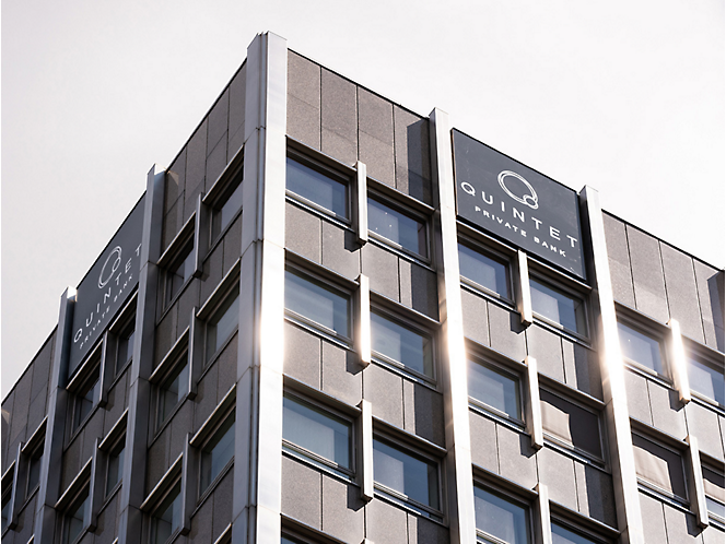 Pilkas modernus biurų pastatas su „quintet“ logotipu ant viršaus, po giedru dangumi, nuo langų atsispindintys saulės spinduliai.
