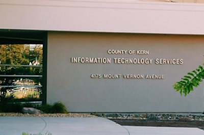 A close-up of a sign on a white wall with black text reading COUNTY OF KERN INFORMATION TECHNOLOGY SERVICES 4175 MOUNT VERNON AVENUE.