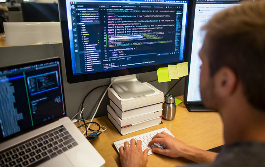 A man in headphones, working in a busy, cluttered office with Visual Studio running on a vertical monitor in the foreground. 