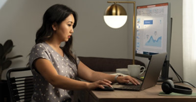 Человек сидит за столом и работает на ноутбуке, подключенном к настольному монитору, где отображается презентация PowerPoint.