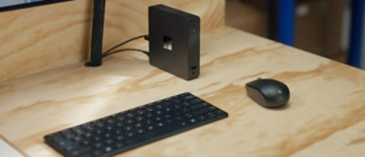 Computer workstation with a keyboard, mouse, and compact desktop device on a wooden desk.