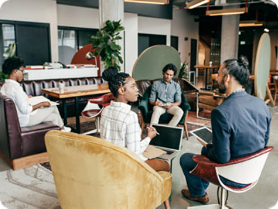 現代のオフィス環境でノート PC を使用してディスカッションに取り組む 4 人の専門家。 2 人はソファに、2 人は肘掛け椅子に座っています。