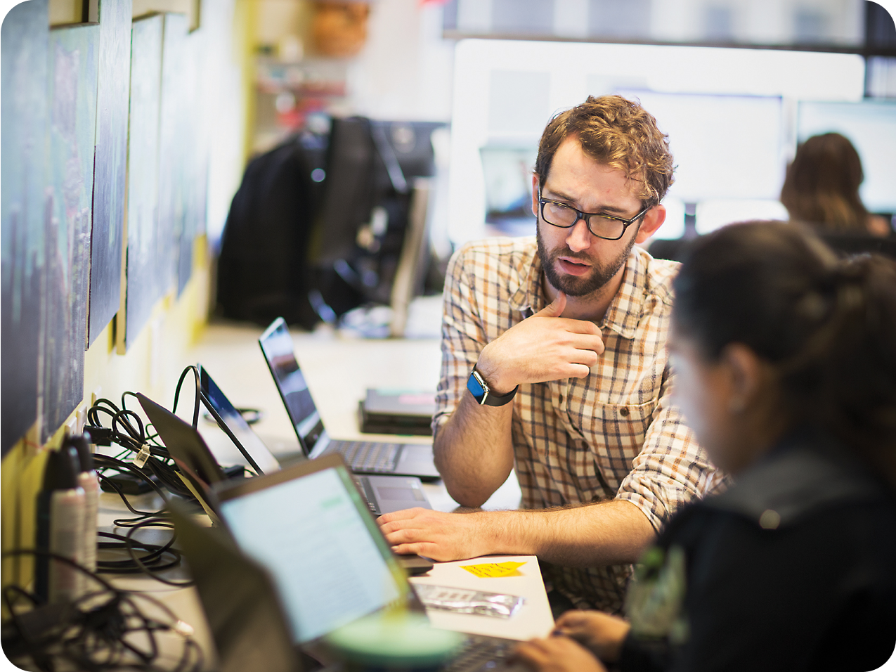 Un homme barbu et portant des lunettes fait des gestes lors d’une discussion avec une collègue, tous deux utilisant des ordinateurs portables dans un environnement de bureau dynamique.