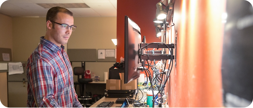Man in glasses and plaid shirt working on multiple monitors in a dimly lit office setting.