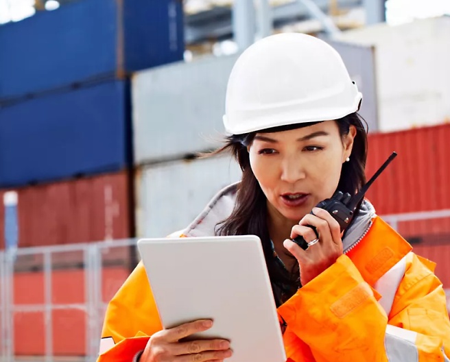A person wearing a hard hat looking at a tablet and using a walkie talkie