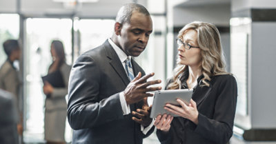 รูปถ่ายของคนสองคนกำลังยืนอยู่ในล็อบบี้อาคารกำลังดูอุปกรณ์แท็บเล็ต มีคนอยู่ในพื้นหลัง