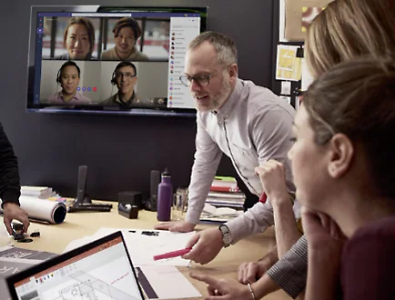 Een groep mensen vergadert in een vergaderzaal en bekijkt bouwkundige blauwdrukken terwijl andere teamleden worden gebeld via een Teams-videogesprek dat wordt weergegeven op een scherm aan de muur achter hen.