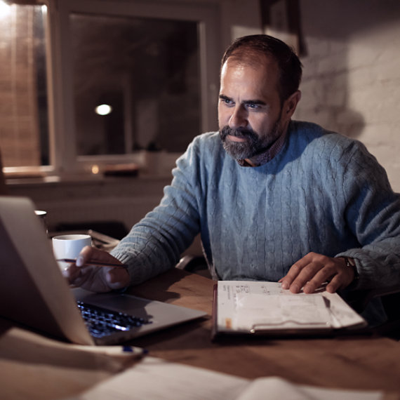 A man working from home in the evening reviews papers on a clip board and updates information on a laptop.