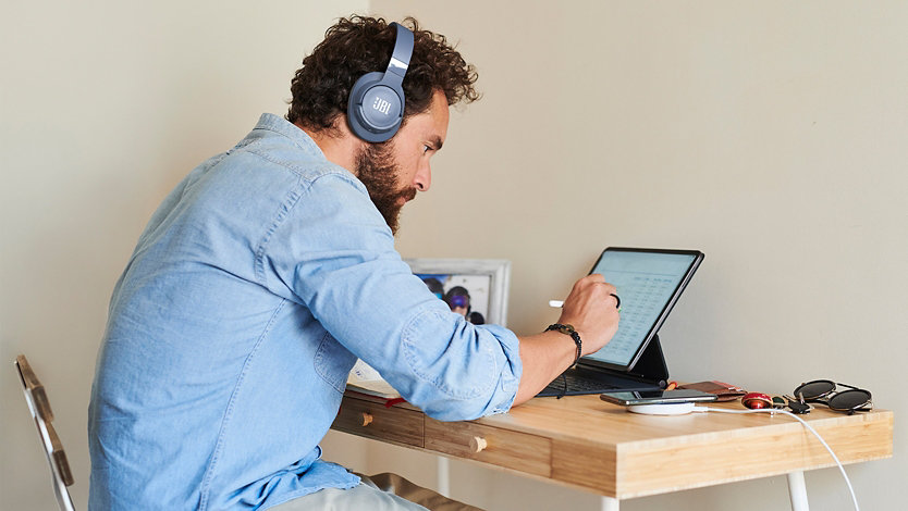 A man wearing the J B L Tune 760 N C Wireless Noise Cancelling Headphones in Blue while working in an office setting.
