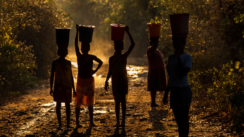 Five women balancing buckets of food and water on their heads.
