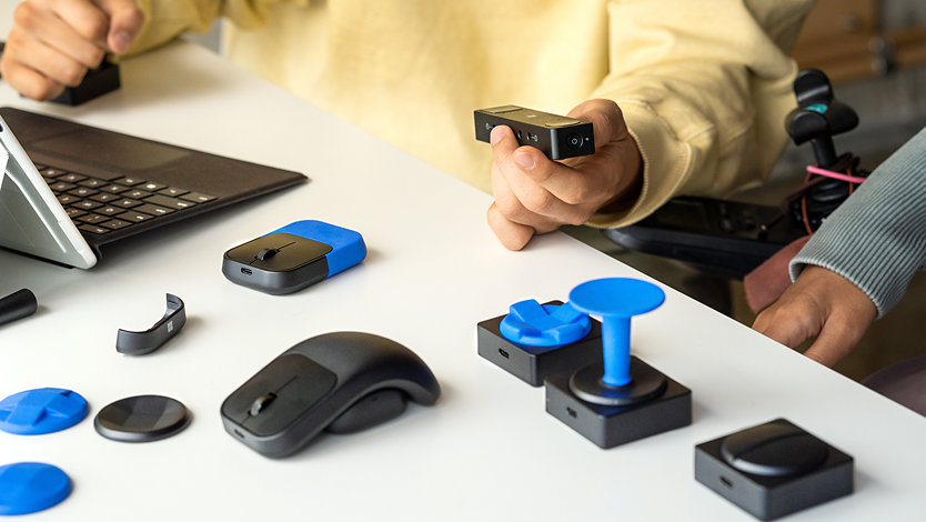 En person bruker Microsoft adaptiv knapp og Microsoft adaptiv mus med Microsoft adaptiv hub i bakgrunnen.