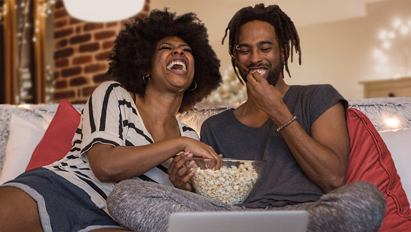 Zwei Menschen lachen und essen Popcorn, während sie fernsehen.