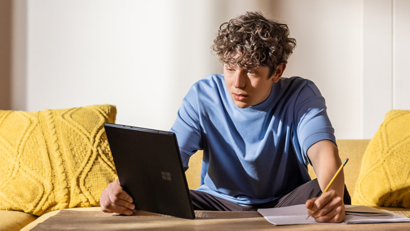 Man seated at a coffee table studying, holding a pencil in one hand and a Surface Pro in the other. 