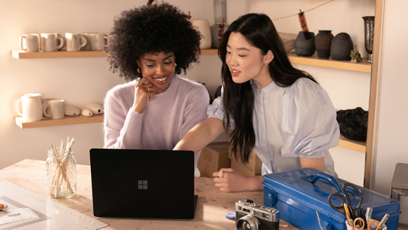 Two people sitting at a table, surrounded by artists’ tools like paint brushes, scissors, and a camera, using a Surface Laptop. 