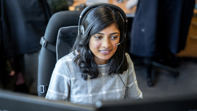 A woman sitting at a desk behind a monitor wearing a headset.