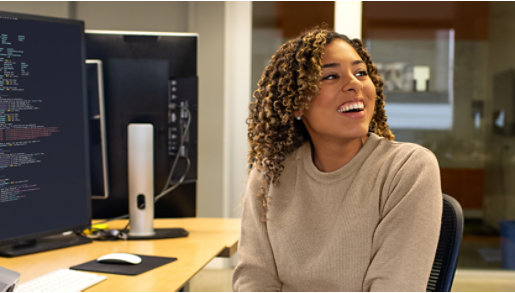 A young Black woman turns away from her computer where she is writing code and smiles.