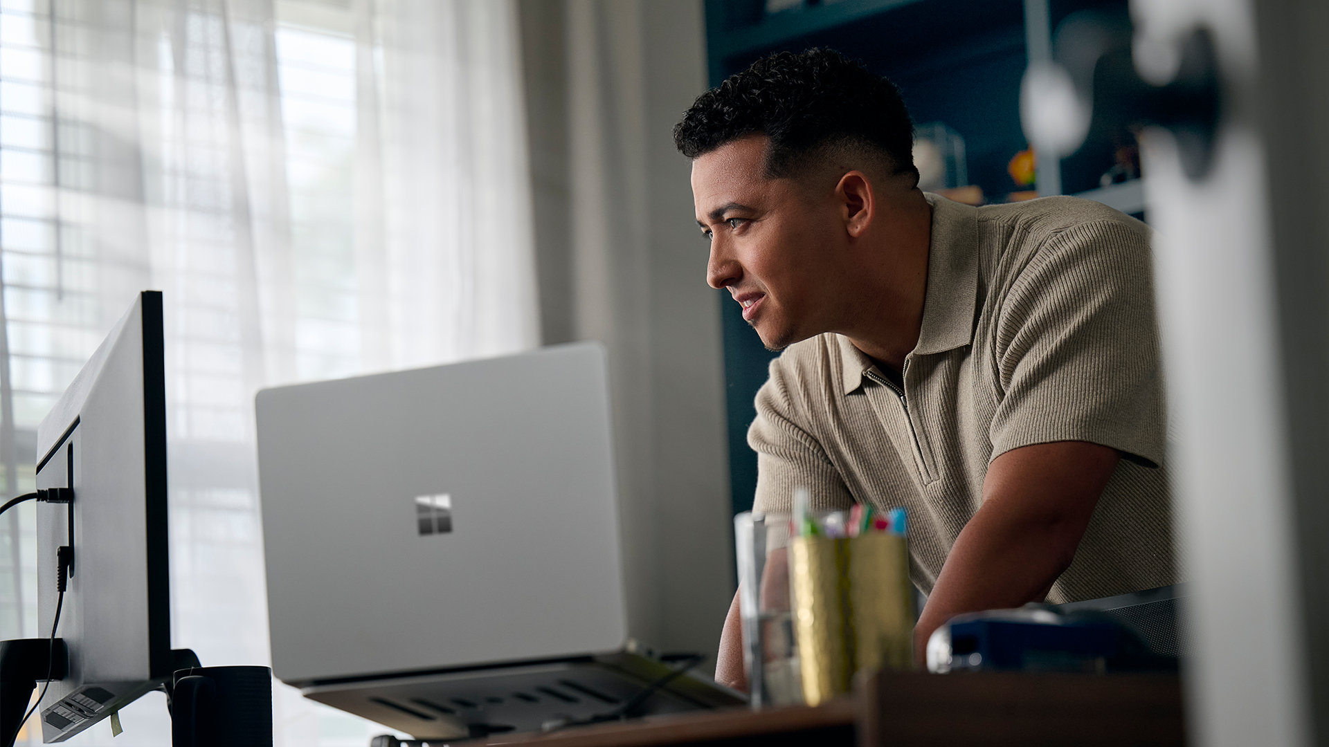 A man leans over a desk working on a Surface Laptop and external monitor. 