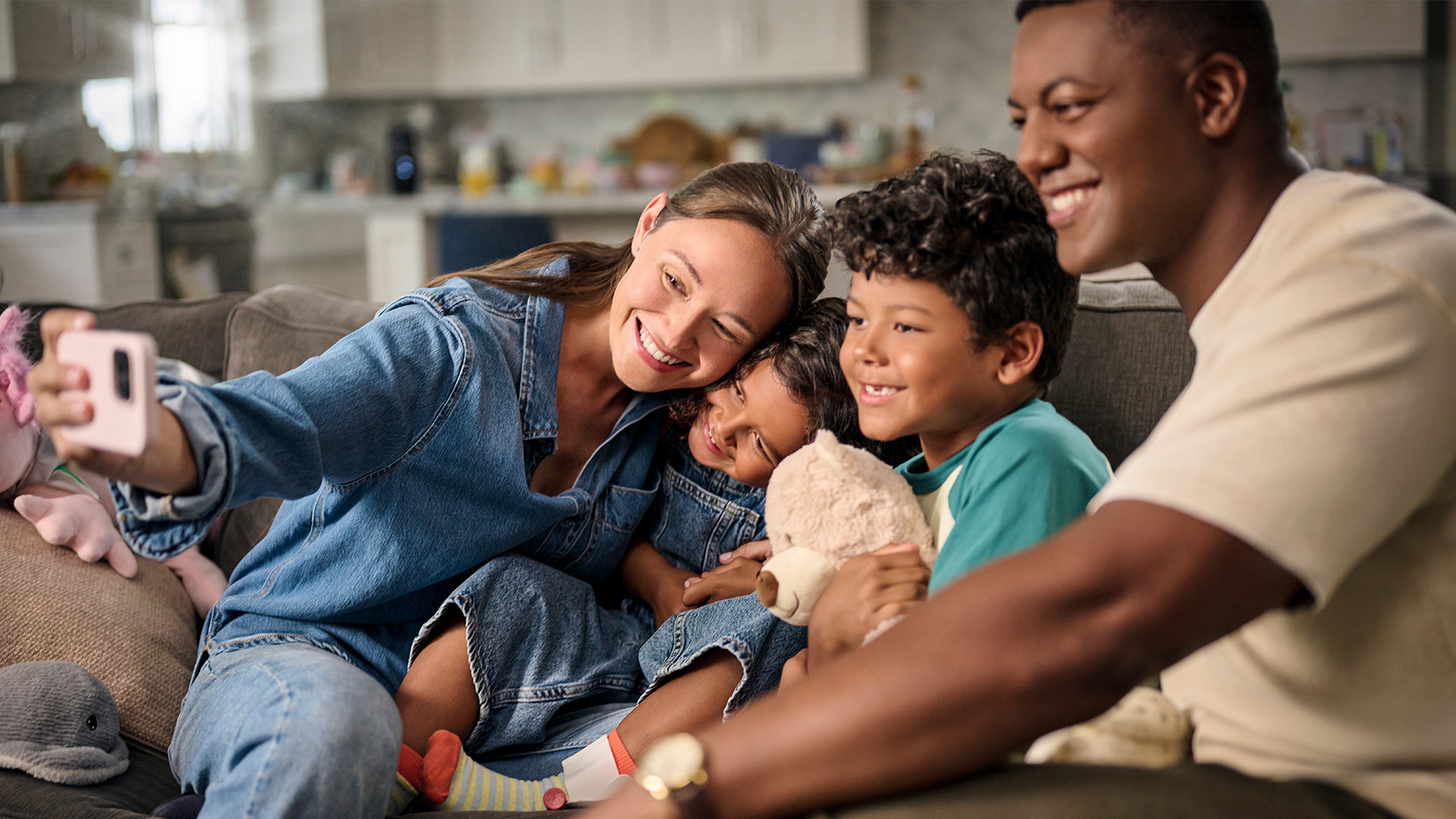 A family of four takes a group selfie while sitting on a living room sofa. 