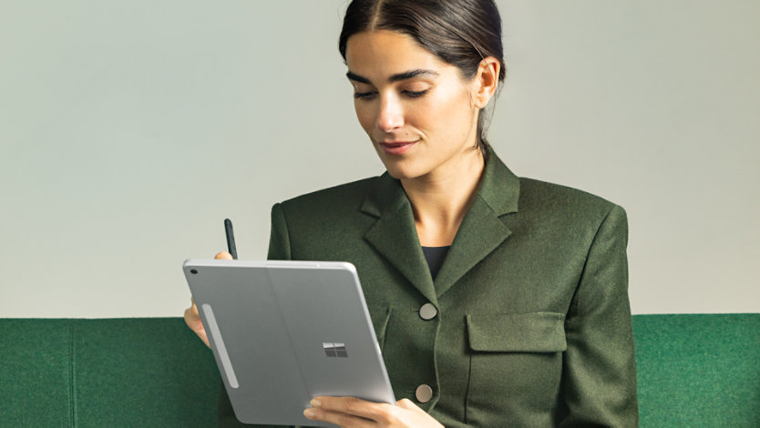 A woman sitting on a green couch, holding a Surface Pro 12 inch Platinum device vertically in tablet mode in her left hand.