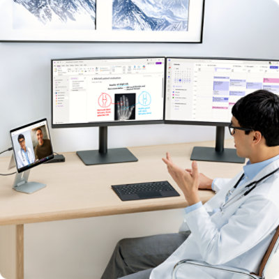 Um médico sentado à secretária numa videochamada, a utilizar a câmara do Surface Pro para Empresas enquanto trabalha em vários monitores..