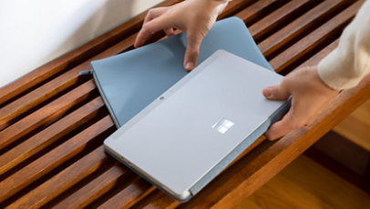 A pair of hands picking up a Surface Go and Ice Blue sleeve.
