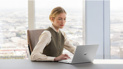 Woman sitting while using a Surface laptop 7 in front of a large plate glass window.