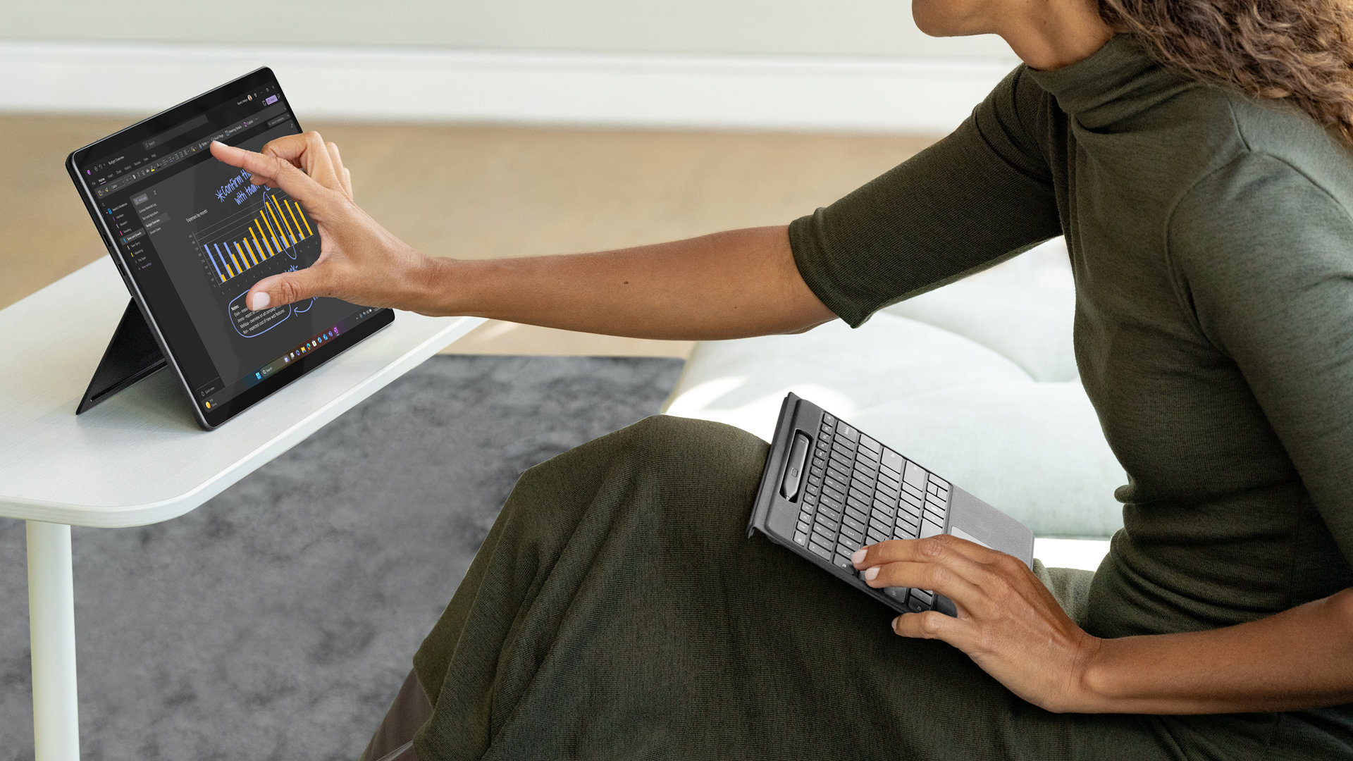 Une personne utilise un clavier Flex pour Surface Pro 13 pouces avec stylet Slim Pen détaché sur ses genoux, tout en interagissant avec l’écran tactile d’un appareil Surface posé sur une table à proximité.