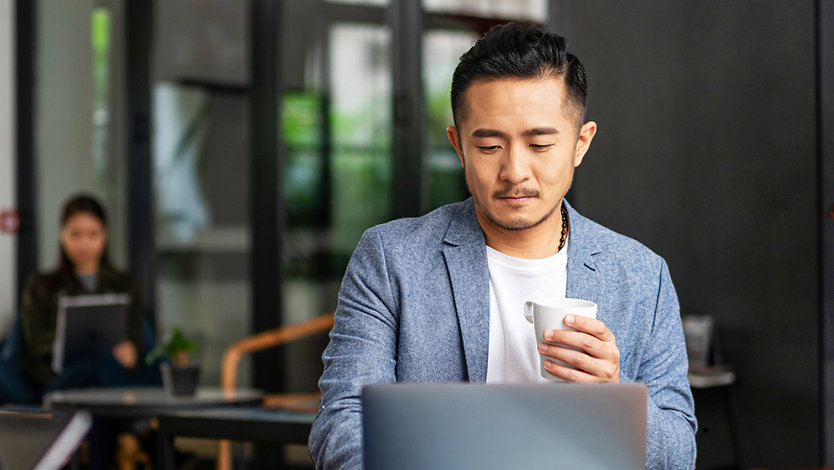 A man in a blazer works on his Surface laptop while holding a coffee while others in the background do the same.