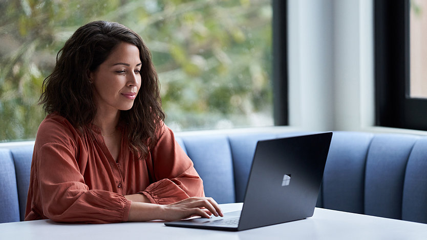 A woman dressed casually works on her Surface laptop while sitting in a padded booth.
