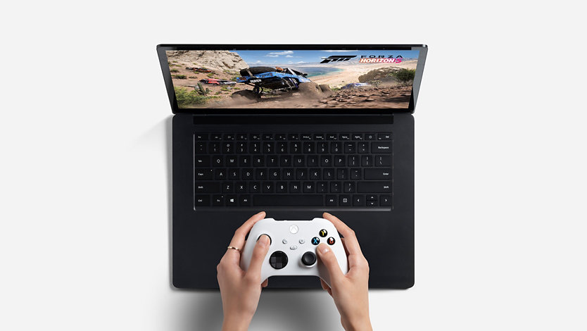 Las manos de una persona jugando con un control de Xbox y su computadora portátil. 