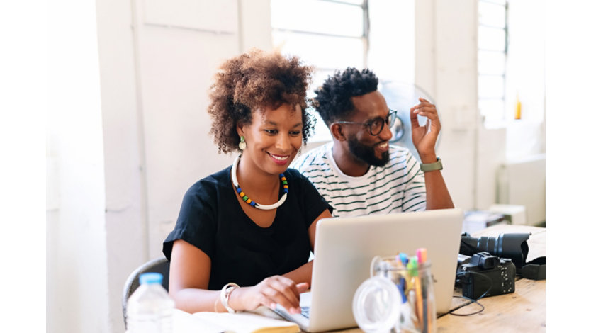 A woman and a man smile while looking at a laptop together at a desk in a bright, modern office. The woman wears a bright, colorful necklace, and a camera sits on the table nearby.